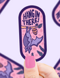 Hand holding a purple bat sticker hanging upside down from a branch with the words Hang in There on pink background