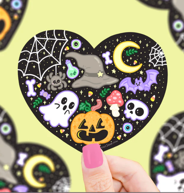 Hand holding a heart-shaped Halloween sticker featuring ghost, pumpkin, bat, spider, moon, and potion bottle designs.