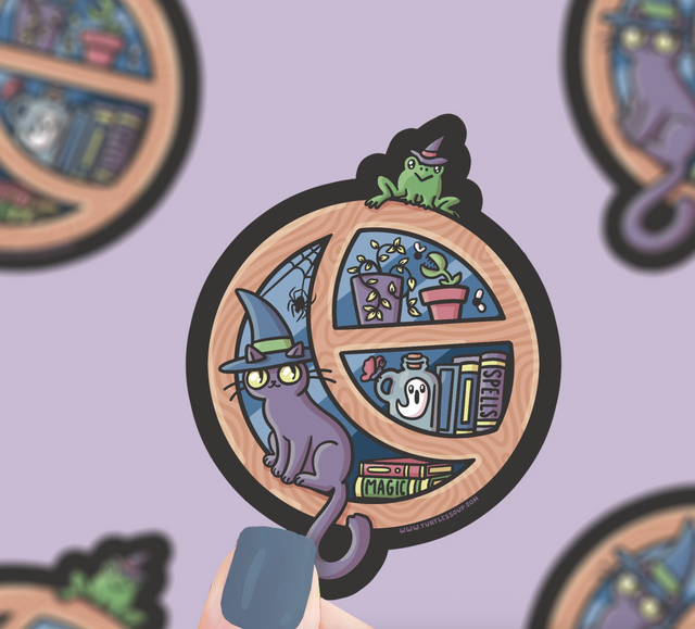 Cute Halloween window sticker showing a witch’s cat, frog, plants, and magical books in a round window frame.