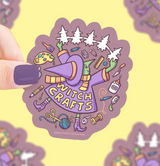 Hand holding a colorful Witch Crafts sticker featuring crafting tools and a witch hat on a purple background