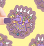 Hand holding a colorful Witch Crafts sticker featuring crafting tools and a witch hat on a purple background