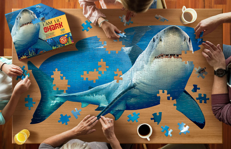 People assembling a 100-piece oversized jigsaw puzzle of a large friendly shark on a wooden table with beverage cups nearby