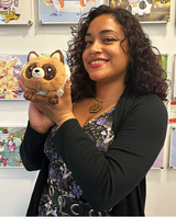 Woman holding a plush Tanuki Snacker toy with a cheerful expression in a colorful art-filled room
