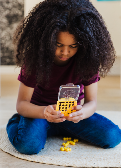 Child focused on solving a yellow IQ Mini puzzle game, sitting cross-legged on a woven mat indoors.