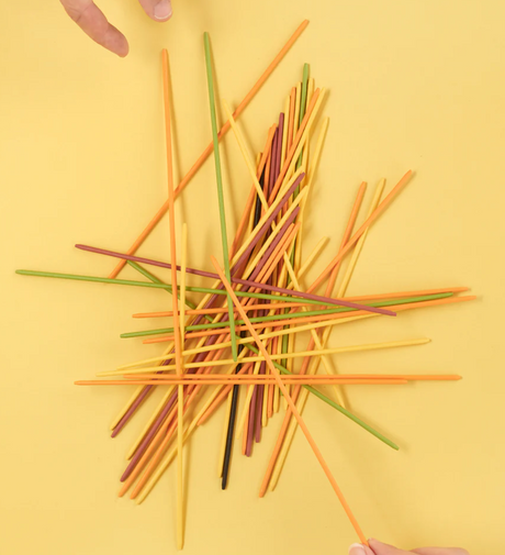 Colorful spaghetti-shaped sticks scattered on a yellow background with hands carefully picking sticks for the game.