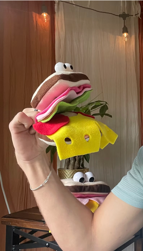 Hand wearing a plush sandwich finger puppet with layers of bread, lettuce, tomato, and cheese in a cozy room setting.