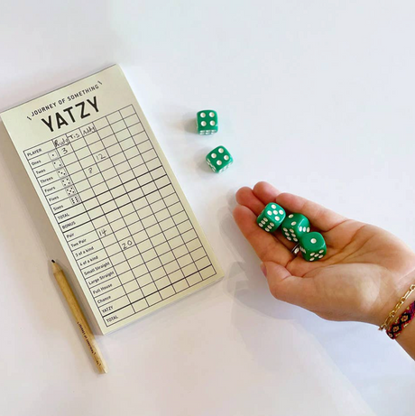 Hand holding green dice next to yatzy score pad and pencil on white surface in bright setting