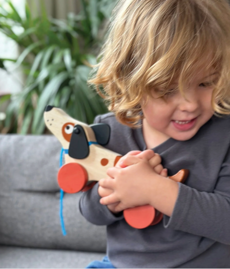 Child smiling and holding a wooden puppy toy with bright red wheels ready for play and adventure indoors