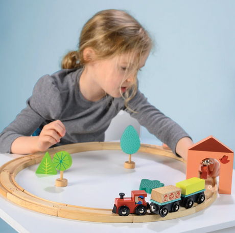 Young child playing with wooden train set, colorful trees, and tunnel on a white surface with a light blue background
