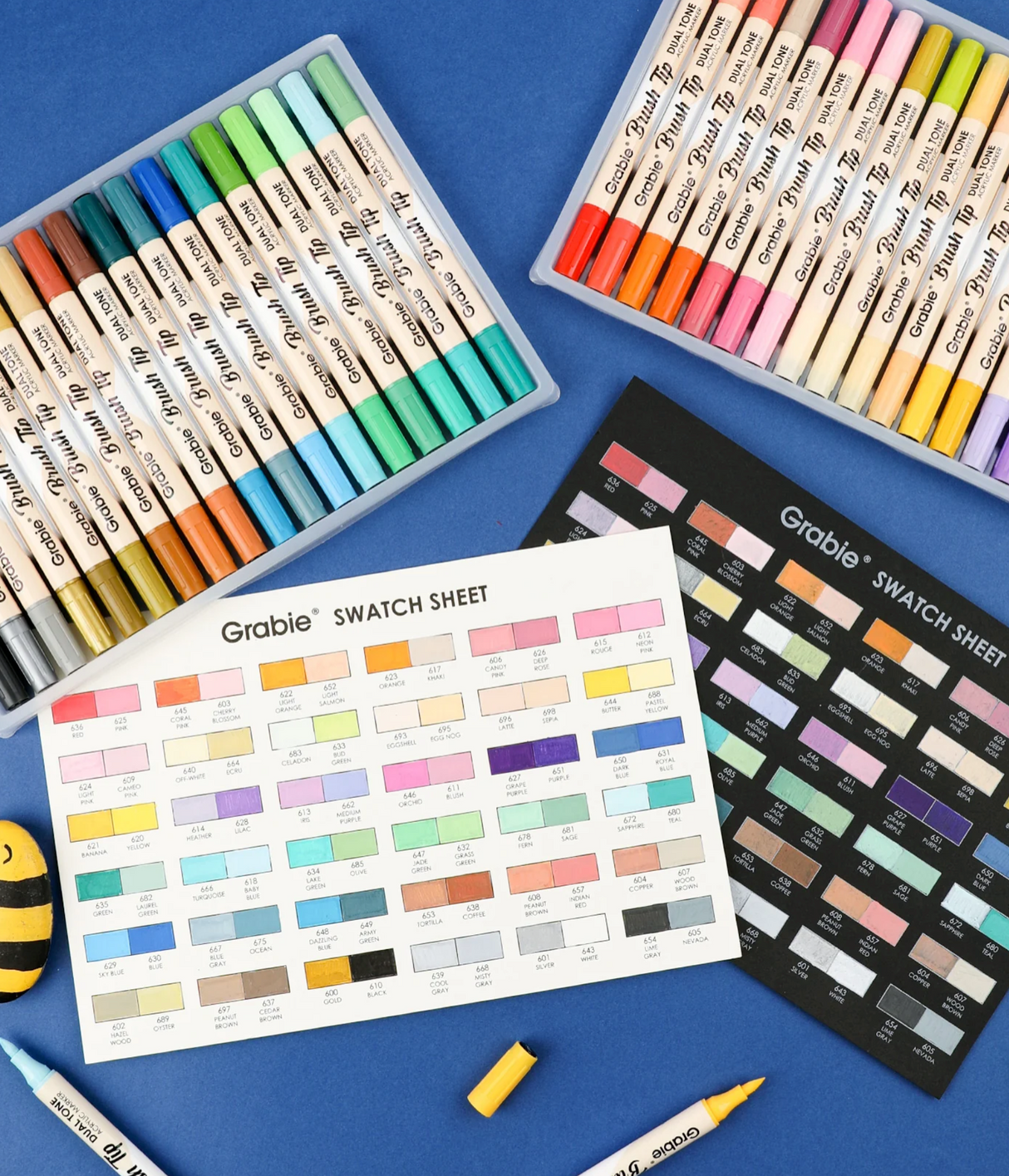 Two sets of duel tone brush tip acrylic paint markers with color swatch sheets on a blue background