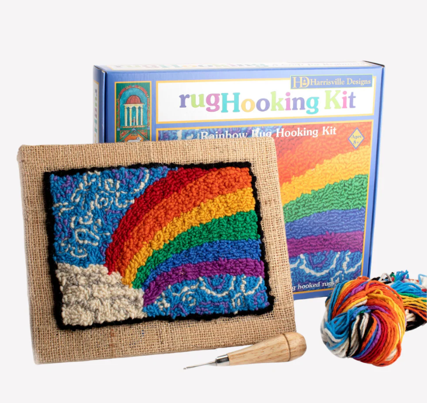 Colorful rainbow design made with wool on burlap canvas beside a rug hooking kit box and multicolored yarn ball