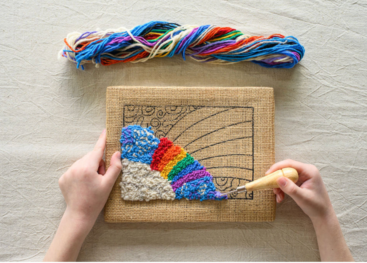 Hands working on colorful rainbow rug hooking on burlap fabric with wool yarns and hooking tool visible