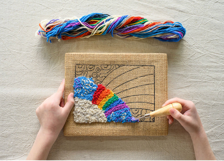 Hands working on colorful rainbow rug hooking on burlap fabric with wool yarns and hooking tool visible