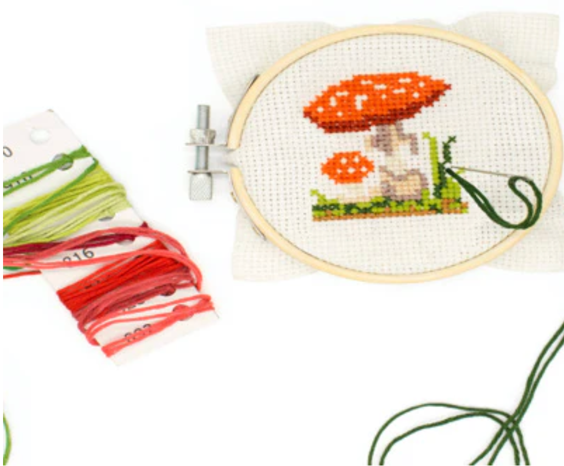Mini cross-stitch embroidery kit with a bamboo hoop frame, colorful threads, and mushroom design in progress.