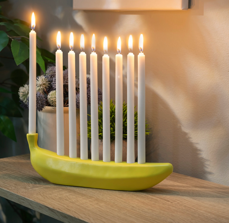 Ceramic menorah shaped like a yellow banana holding nine lit white candles on a wooden surface.