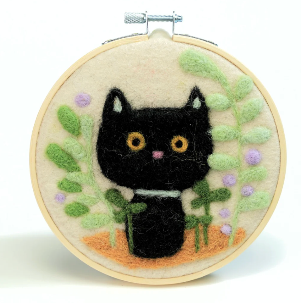 Needle felted black cat surrounded by green leaves and purple flowers in wooden embroidery hoop.