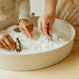 Two children playing with small penguin figures in a large bowl filled with textured sensory snow material