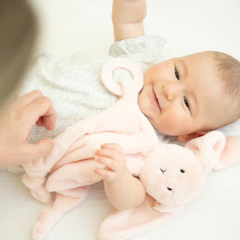 Smiling baby lying down holding a soft pink bunny teether buddy with a plush body and teething ring attachment