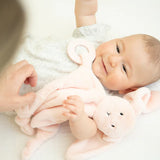 Smiling baby lying down holding a soft pink bunny teether buddy with a plush body and teething ring attachment