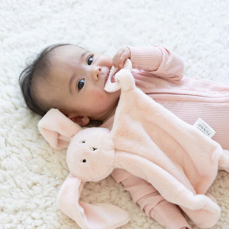 Infant lying on soft blanket holding and chewing on a soft plush bunny teether buddy toy