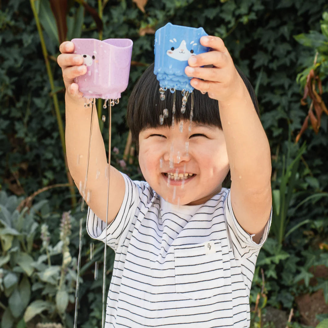 Child playing outdoors holding two colorful animal cups pouring water with joyful expression