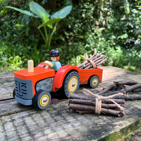 Farmyard Tractor wooden toy with removable trailer, driver figure, and bundled sticks on rustic outdoor table.