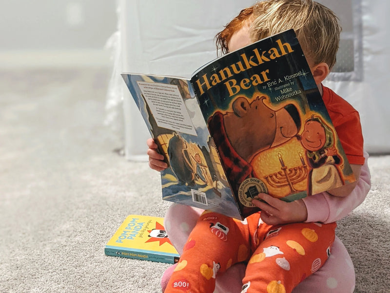 Young child sitting on carpet reading the book Hanukkah Bear with another book nearby