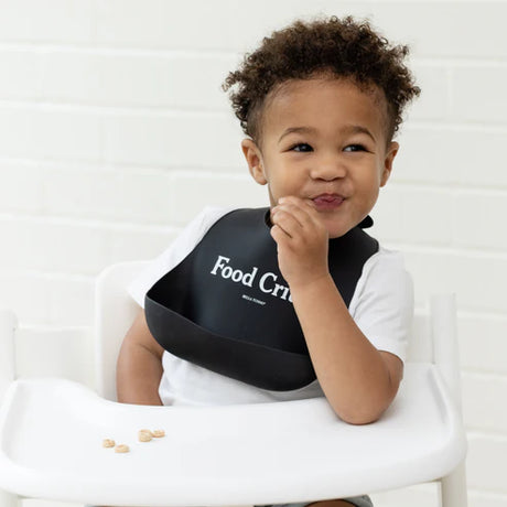 Baby wearing Wonder Bib | Food Critic silicone bib with catch pocket sitting in high chair, smiling and eating snacks.