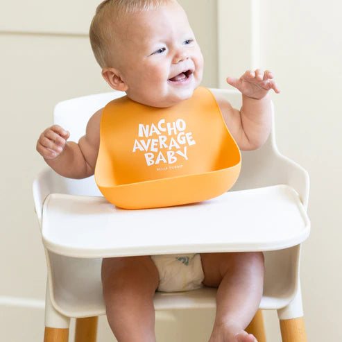Smiling baby wearing orange Wonder Bib with catch pocket sitting in white high chair ready for mealtime