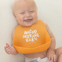 Smiling baby wearing orange bib with catch pocket and "Nacho Average Baby" text for clean mealtime fun