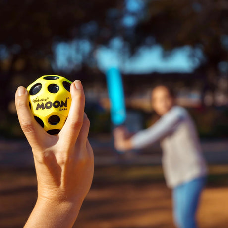 Person holding yellow Moon ball ready to hit with blue Waboba Land Cracket bat in background outdoors, Waboba Land Cracket.