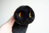 Close-up of a black Wooly Bear Caterpillar weighted plush with soft faux fur and embroidered yellow eyes held in hand