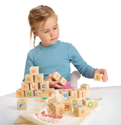 Child playing with colorful wooden alphabet blocks stacking and spelling on a white table with a drawstring bag nearby