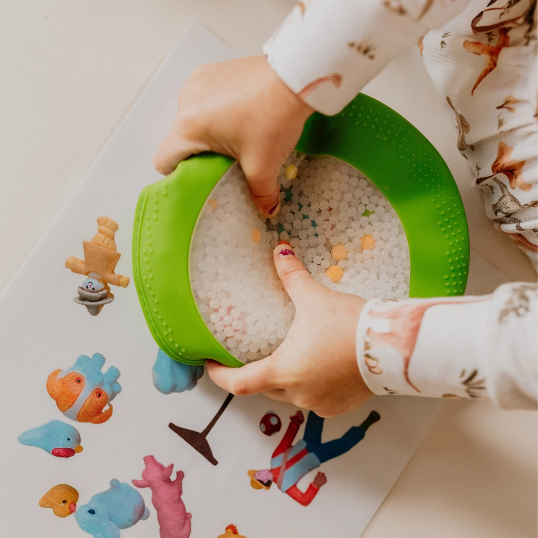 Child's hands squeezing a green sensory toy filled with white beads over colorful animal stickers on a white surface