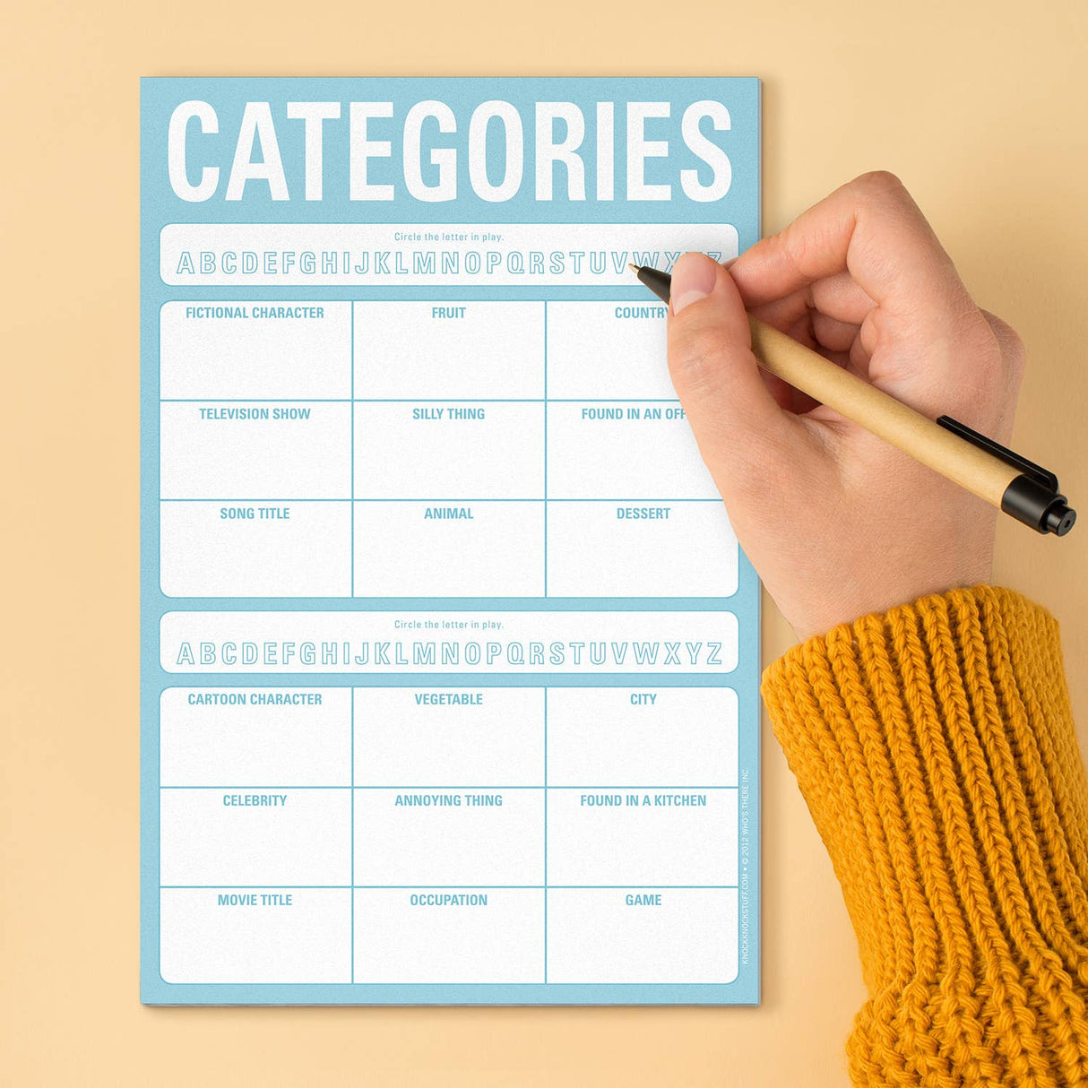 Hand holding pencil filling out a categories game sheet from the On-the-Go Game Pad on a beige surface