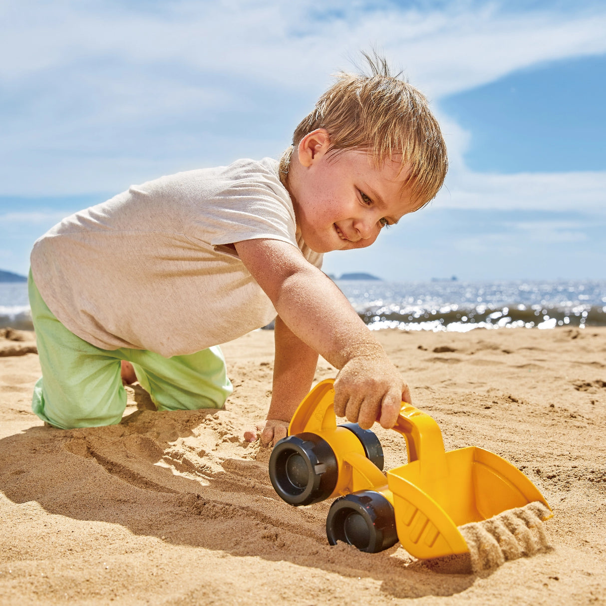 Young boy playing with a yellow toy excavator digging sand on a sunny beach near the water
