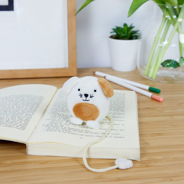 Fluffy white and brown animal-shaped bookmark resting on an open book on a wooden surface beside plants and pens