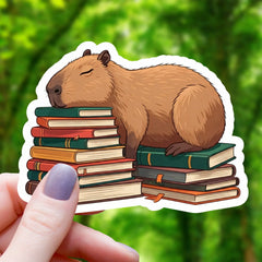 Capybara Sleeping on Books Sticker featuring a sleepy capybara resting on a colorful stack of books