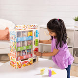 Young girl playing with a wooden vending machine toy filled with colorful pretend snacks and drinks at a table