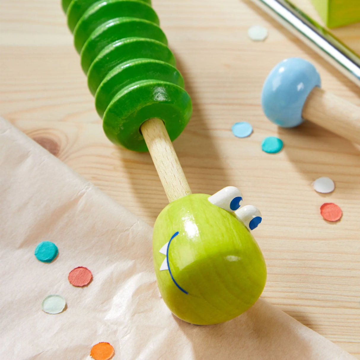 Close-up of a wooden slide whistle shaped like a smiling green crocodile on a light wooden surface with colorful confetti pieces