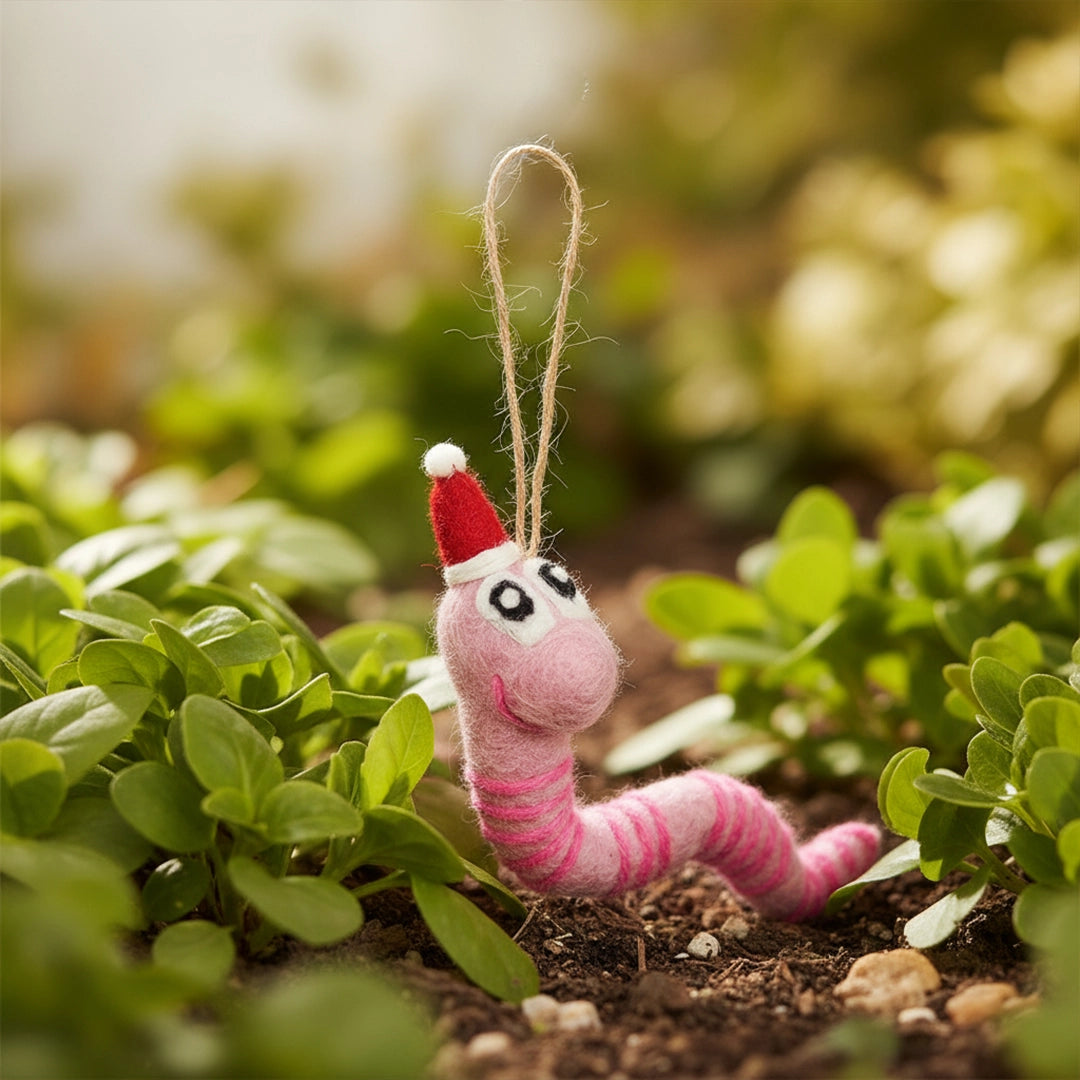 Pink felt earthworm ornament wearing a red Santa hat with big eyes hanging among green plants outdoors