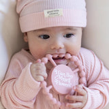Baby wearing pink ribbed hat and outfit holding a pink teething ring with motivational white text