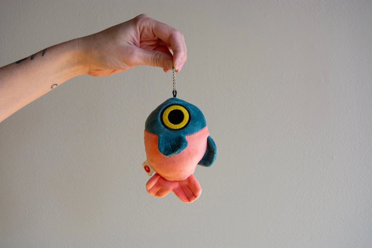 Hand holding a soft squishy mochi fabric fish-shaped plush keychain with a chain against a plain background