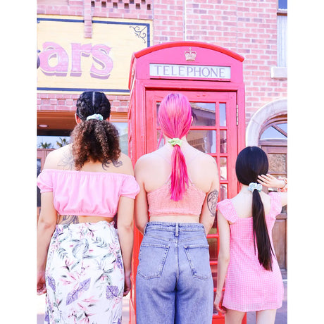 Three women with different hairstyles wearing the Banana Hair Scrunchie standing in front of a red phone booth