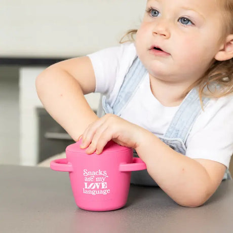 Toddler using a pink Happy Snacker | Snacks Are My Love Language silicone snack cup with easy grip handles and snap tight lid.
