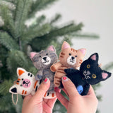 Handmade felt cat ornaments in various colors holding small fish, crafted from 100% wool with detailed needle felting.