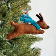 Handmade felt jumping dog ornament with blue scarf hanging on a Christmas tree branch.
