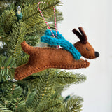 Handmade felt jumping dog ornament with blue scarf hanging on a Christmas tree branch.