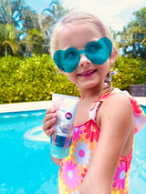 Young girl wearing blue heart-shaped sunglasses and floral swimsuit holding a sunscreen tube by a sunny poolside