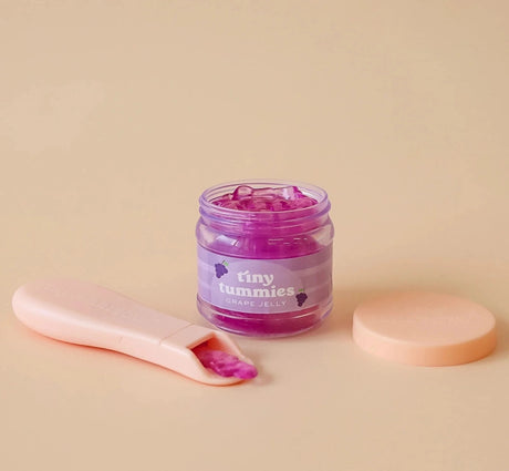 Small jar of grape jelly food with a pastel pink spoon and lid on a soft beige background for dolls.